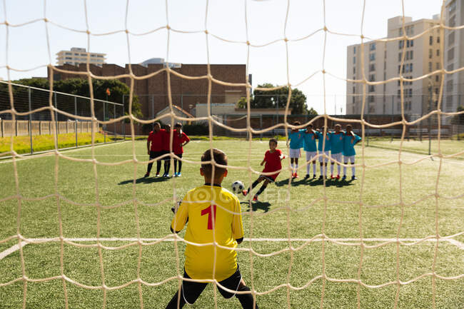 Rear view of a Caucasian boy in goal while a team takes penalty shots against him, during a match between two multi-ethnic teams of boy soccer players wearing their team strips in action on a football pitch — Stock Photo