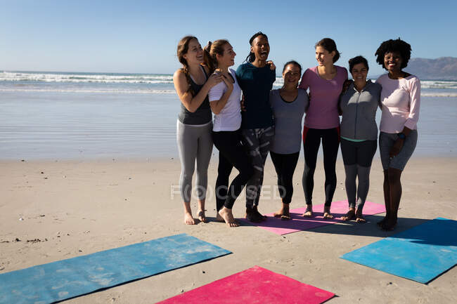 Vue de face d'un groupe multi-ethnique d'amies profitant du temps passé ensemble sur une plage par une journée ensoleillée, debout derrière les tapis de yoga, portant des vêtements de sport, souriant, embrassant. — Photo de stock