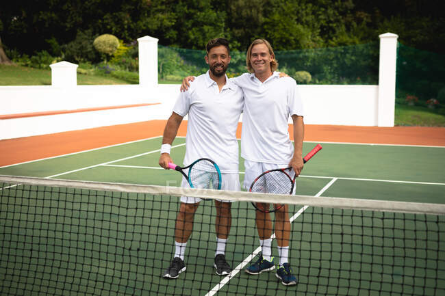 Портрет кавказца и смешанной расы мужчин в теннисных белках, проводящих время на площадке вместе, играющих в теннис в солнечный день, обнимающихся, держащих теннисные ракетки, смотрящих в камеру и улыбающихся — стоковое фото