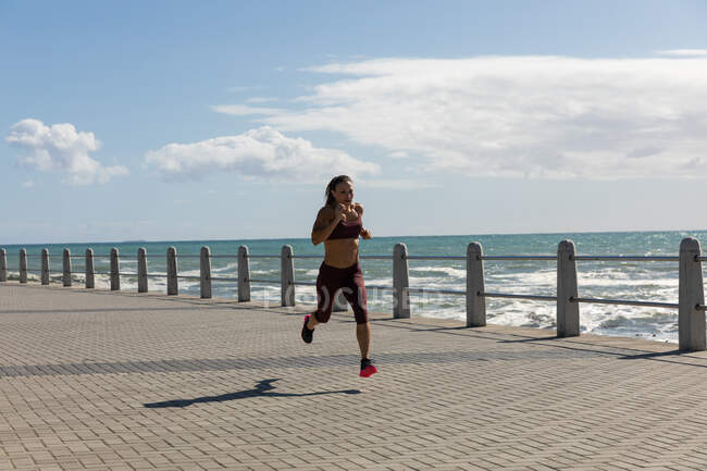 Vista lateral de una mujer atlética caucásica con el pelo largo y oscuro haciendo ejercicio en un paseo marítimo en un día soleado con cielo azul, corriendo. - foto de stock