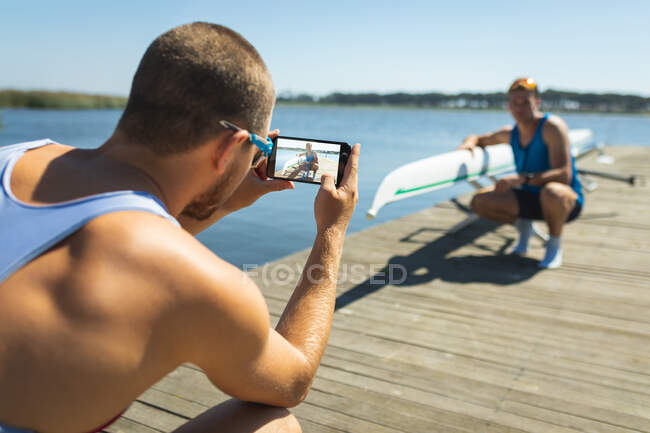 Vista trasera de un remo macho caucásico tomando fotos con el smartphone de su amigo posando junto a un bote de remos en un embarcadero en el río - foto de stock