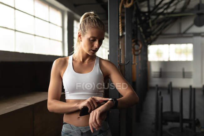 Vorderansicht einer athletischen kaukasischen Frau in Sportkleidung beim Crosstraining in einem Fitnessstudio, stehend und mit Hilfe einer Fitness-App auf die Smartwatch blickend — Stockfoto