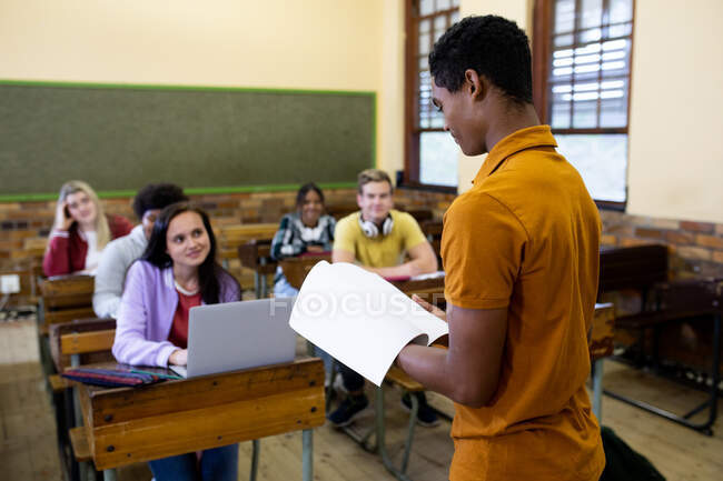 Visão lateral de um adolescente afro-americano estudante do ensino médio lendo para uma classe multi-étnica de adolescentes sentados em mesas em uma sala de aula da escola ouvindo e concentrando — Fotografia de Stock