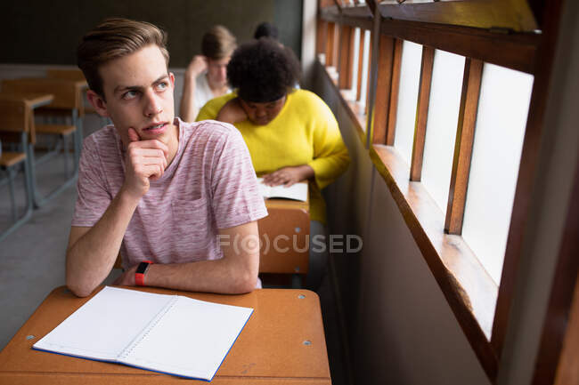 Vista frontal de un adolescente caucásico en un aula de la escuela sentado en el escritorio, concentrándose y mirando por la ventana, con compañeros de clase adolescentes masculinos y femeninos sentados en escritorios trabajando en el fondo - foto de stock