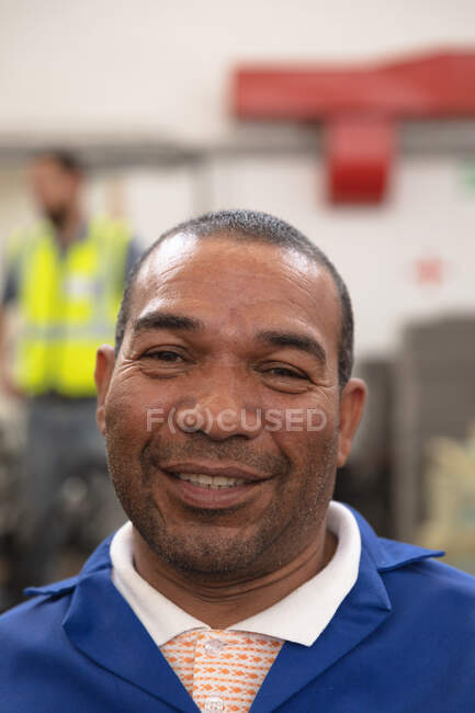 Retrato de um trabalhador mestiço vestindo um vestuário de trabalho, em um armazém de armazenamento em uma fábrica fazendo cadeiras de rodas, olhando para a câmera e sorrindo — Fotografia de Stock