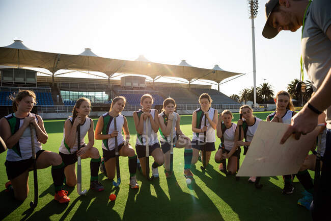 Front view of a group of female Caucasian field hockey players preparing before a game, kneeling in front of their Caucasian male coach, on a hockey pitch, who gives them game plan, on a sunny day — Stock Photo