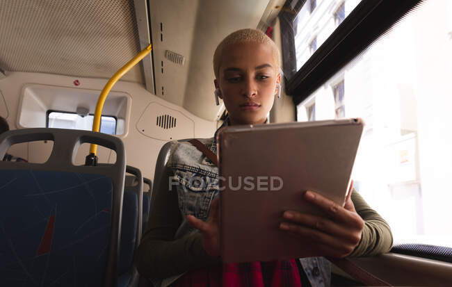 Femme alternative de race mixte avec les cheveux blonds courts dehors et environ dans la ville, assis sur un bus portant des écouteurs sans fil et utilisant un ordinateur tablette. Nomade numérique urbain en déplacement. — Photo de stock