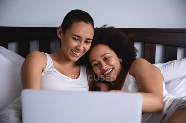 Вид спереду крупним планом змішаної раси жіноча пара розслабляється вдома в спальні, сидячи в ліжку, використовуючи ноутбук разом і посміхаючись — стокове фото
