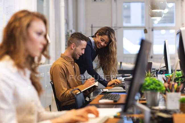 Caucasian male business creative working in a casual modern office, sitting at a desk and smiling, taking notes with female colleagues working next to him — Stock Photo