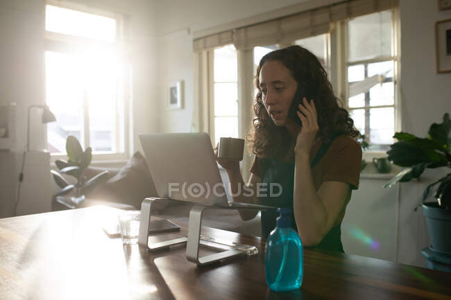 Mujer caucásica pasando tiempo en casa, sentada junto a su escritorio y trabajando, hablando en un smartphone y usando su portátil. Distanciamiento social y autoaislamiento en cuarentena. - foto de stock
