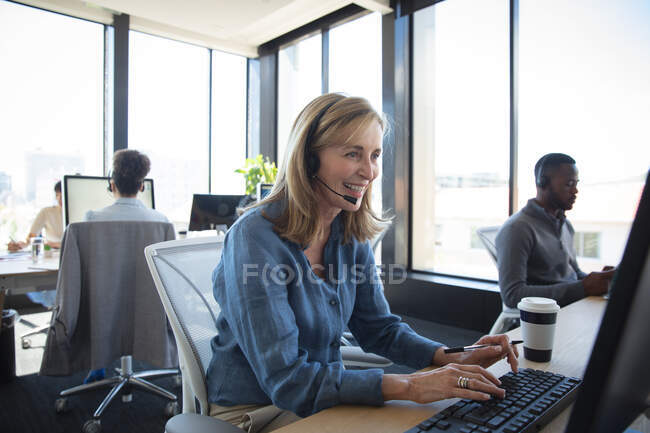 Una donna d'affari caucasica che lavora in un ufficio moderno, seduta alla scrivania, usando un computer portatile, indossando cuffie e parlando, con i suoi colleghi che lavorano sullo sfondo — Foto stock