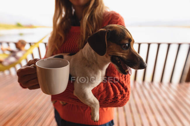 Vue de face d'une femme caucasienne s'amusant lors d'un voyage à la montagne, debout sur un balcon dans une cabine, tenant une tasse de café, et un chiot — Photo de stock
