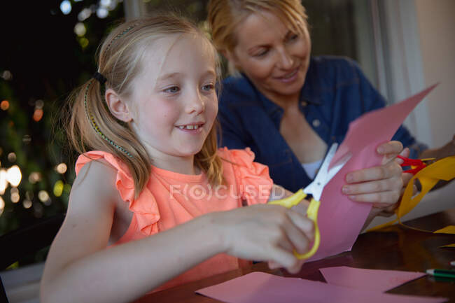 Vista lateral de una mujer caucásica disfrutando de tiempo en familia con su hija en casa juntos, sentada en una mesa en la sala de estar y cortando papeles con tijeras - foto de stock