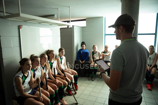 Seitenansicht eines kaukasischen Hockeytrainers, der mit einer Gruppe kaukasischer Hockeyspielerinnen interagiert, in einer Umkleidekabine sitzt und ein digitales Tablet in der Hand hält — Stockfoto