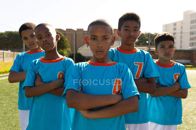 Front view of a multi-ethnic group of boy soccer players wearing their team strip, standing on a playing field in the sun, looking straight to camera with arms crossed — Stock Photo