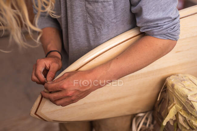 Mittelteil eines männlichen Surfbrettmachers mit langen blonden Haaren, in seinem Studio, poliert eine Kante des Surfbretts aus Holz. — Stockfoto