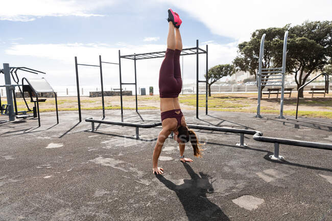 Vista lateral de una mujer atlética caucásica con pelo largo y oscuro haciendo ejercicio en un gimnasio al aire libre durante el día, haciendo handstand. - foto de stock