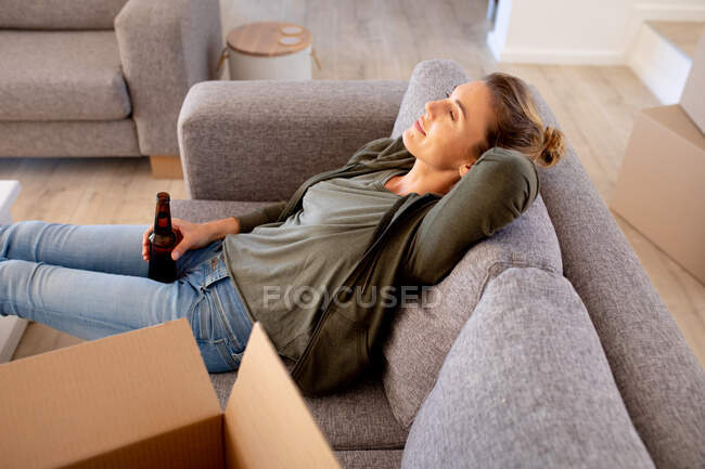 Mujer caucásica pasar tiempo en casa auto aislamiento y distanciamiento social en cuarentena de bloqueo durante coronavirus covid 19 epidemia, tomando un descanso mientras hace bricolaje, descansando en un sofá y bebiendo una cerveza. - foto de stock