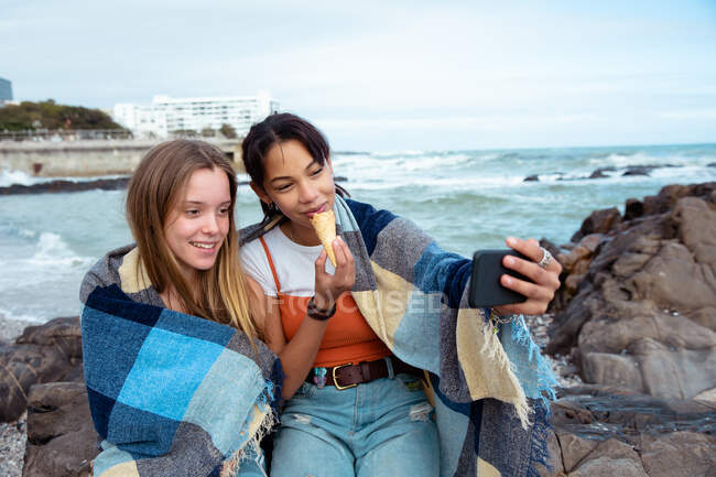Vista frontal de um caucasiano e uma raça mista meninas desfrutando de tempo pendurado juntos em um dia ensolarado, comer sorvete, sentado em uma rocha na praia coberta com o cobertor . — Fotografia de Stock