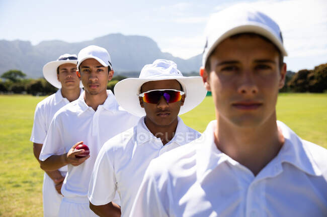 Front view close up of a teenage multi-ethnic male cricket team wearing whites, standing on the pitch together in a row looking straight to camera. — Stock Photo