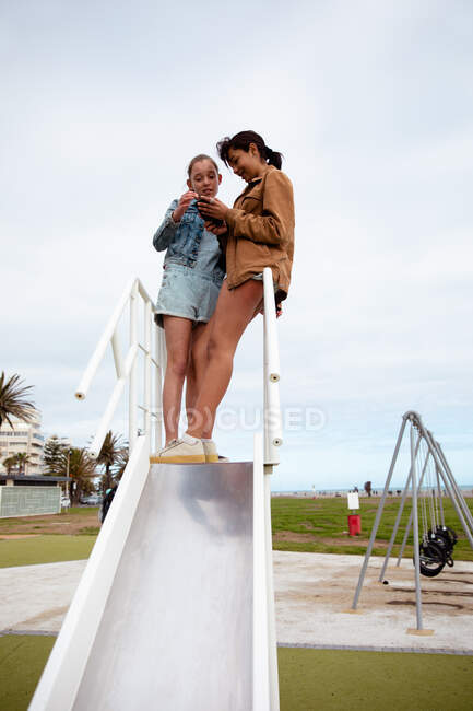 Низькокутний вид на кавказьку і змішану расу дівчат, які проводять час разом у сонячний день, стоять разом на вершині слайда, дівчина тримає свій смартфон, показуючи його своєму другові. — стокове фото