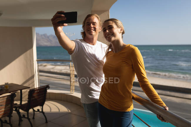 Coppia caucasica in piedi su un balcone, abbracciando e prendendo un selfie insieme. Distanziamento sociale e autoisolamento in quarantena. — Foto stock