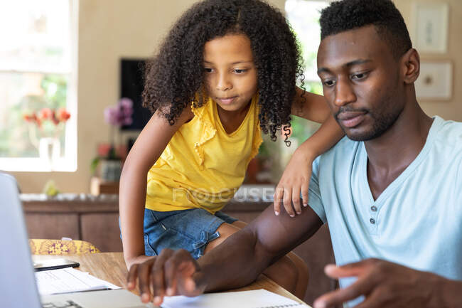 Chica afroamericana con una blusa amarilla, distanciamiento social en casa durante el bloqueo de cuarentena, pasar tiempo con su padre usando una computadora portátil. - foto de stock