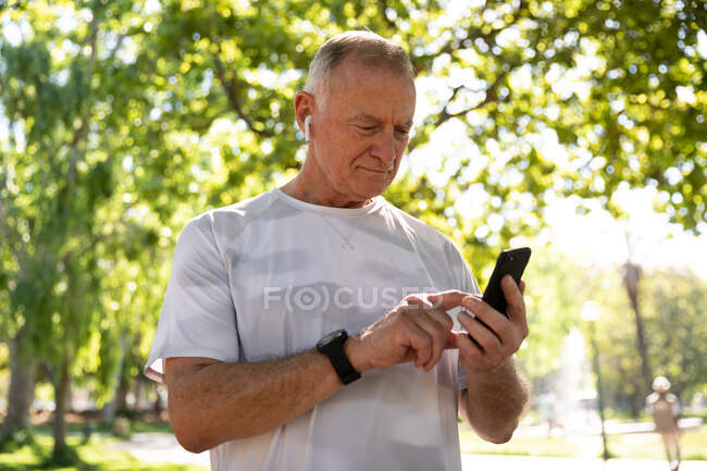 Vista frontal de un hombre caucásico mayor maduro haciendo ejercicio en un parque en un día soleado, usando un teléfono inteligente, preparándose para hacer ejercicio - foto de stock