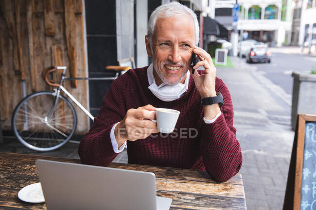 Hombre caucásico mayor sentado en una mesa en una terraza de café, con una máscara facial contra el coronavirus, covid 19, tomando café y usando un teléfono inteligente. - foto de stock