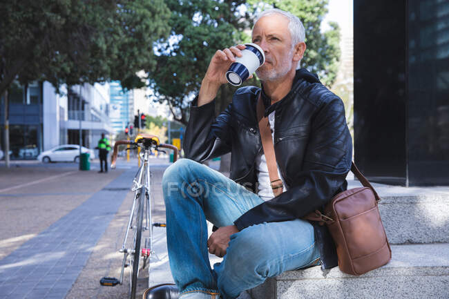 Hombre caucásico mayor, vestido con ropa casual, fuera y alrededor de las calles de la ciudad durante el día, sentado en las escaleras y sosteniendo una taza de café para llevar. - foto de stock