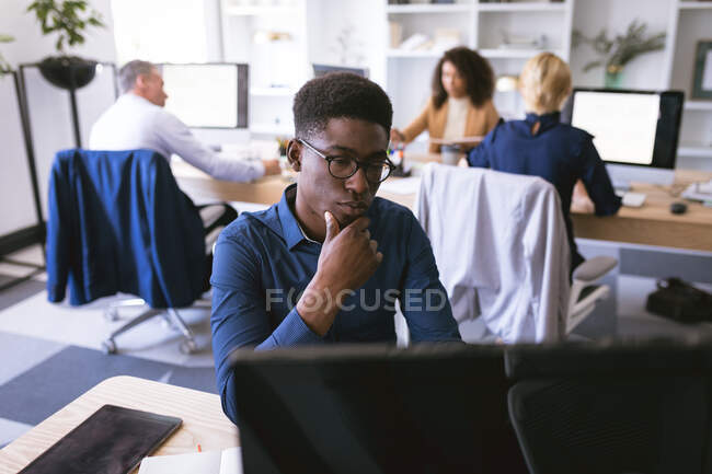 Афроамериканский бизнесмен, работающий в современном офисе, сидящий за столом и пользующийся компьютером, со своими коллегами по бизнесу, работающими на заднем плане — стоковое фото