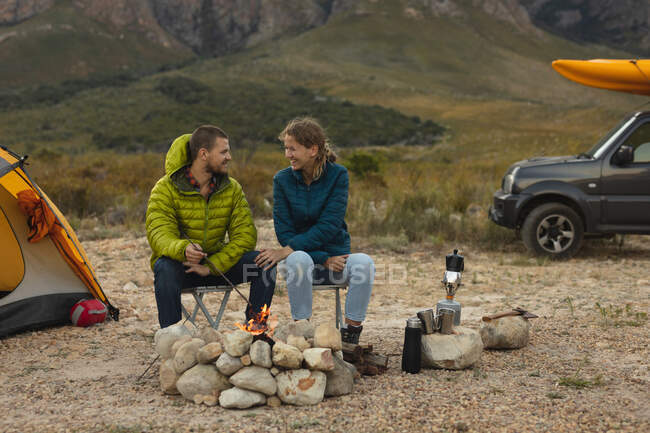 Vorderansicht eines kaukasischen Paares, das sich bei einem Ausflug in die Berge amüsiert, am Lagerfeuer sitzt, einem Feuer zusieht, einander anschaut, eine Frau hält ihre Hand auf einem Männerbein — Stockfoto