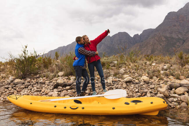 Vista lateral de um casal caucasiano se divertindo em uma viagem às montanhas, abraçando e tirando uma selfie depois de colocar um caiaque na água — Fotografia de Stock