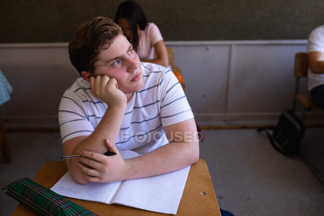 Vue de face en angle élevé d'un adolescent caucasien assis à un bureau dans une classe d'école regardant ailleurs, pensant, camarades de classe assis à des bureaux en arrière-plan — Photo de stock
