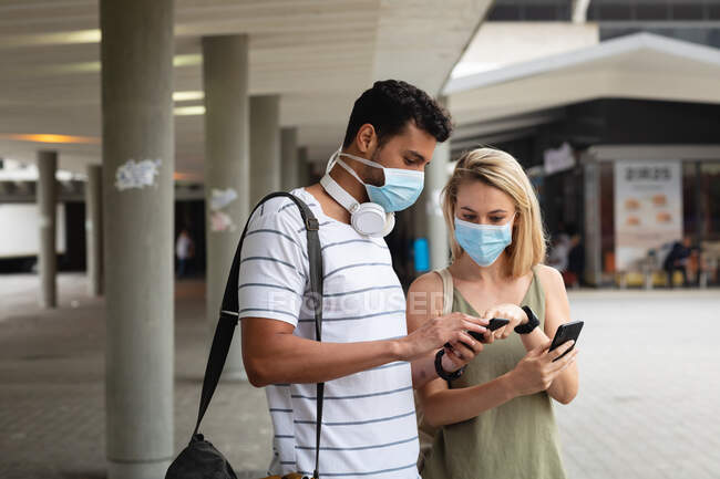 Vista frontal de una pareja caucásica por las calles de la ciudad durante el día, con máscaras faciales contra la contaminación del aire y covid19 coronavirus, utilizando sus teléfonos inteligentes. - foto de stock