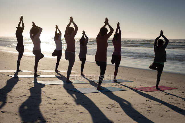 Rear view of a multi-ethnic group of female friends enjoying exercising on a beach on a sunny day, practicing yoga, standing in yoga position. — Stock Photo
