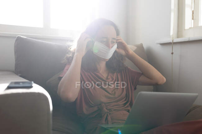 Mulher caucasiana passar o tempo em casa, vestindo um vestido rosa, sentado em um sofá e segurando seu computador portátil, colocando uma máscara facial contra coronavírus, covid 19
. — Fotografia de Stock