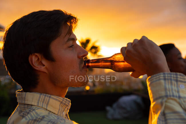 Vista lateral de un hombre caucásico colgando en una azotea con un cielo al atardecer, sosteniendo una botella de cerveza y bebiendo - foto de stock