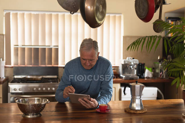 Передній вид на старшого кавказького чоловіка, який відпочиває вдома, сидячи за прилавком на кухні за допомогою планшетного комп 