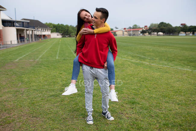 Vista frontal de una pareja de estudiantes de secundaria caucásicos de sexo femenino y masculino pasando el rato, de pie en sus terrenos de la escuela, el chico apoyando a la chica - foto de stock