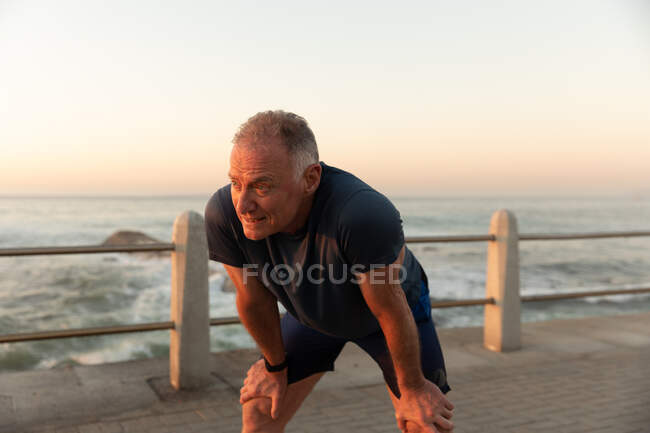 Vista frontal de um homem maduro caucasiano sênior trabalhando em um passeio no dia ensolarado, apoiando-se de joelhos, fazendo uma pausa junto ao mar — Fotografia de Stock