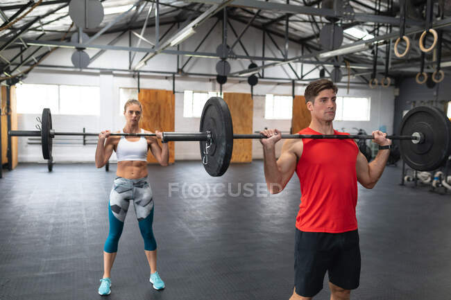 Vista frontal de un atlético hombre y mujer caucásicos con ropa deportiva cruzando el entrenamiento en un gimnasio, de pie y entrenamiento con pesas con barras, levantándolas y sosteniéndolas a nivel del pecho - foto de stock
