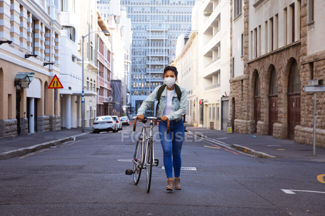 Vue de face d'une femme métisse aux cheveux foncés dans les rues de la ville pendant la journée, portant un masque facial contre la pollution atmosphérique et le coronavirus, marchant avec son vélo avec des bâtiments en arrière-plan. — Photo de stock