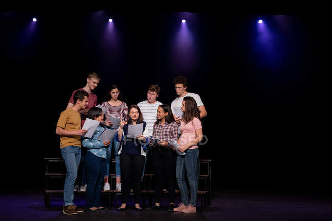 Vista frontal de un grupo multiétnico de coristas adolescentes masculinos y femeninos sosteniendo partituras y cantando de pie en el escenario de un teatro escolar durante los ensayos para una actuación - foto de stock