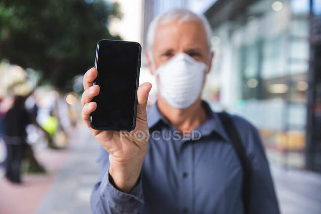 Homem caucasiano sênior nas ruas da cidade durante o dia, usando uma máscara facial contra coronavírus, vívido 19 e mostrando um smartphone . — Fotografia de Stock