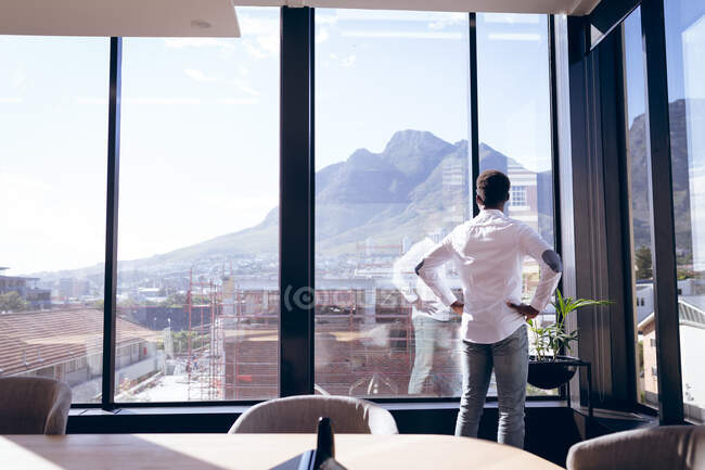 Афроамериканський бізнесмен, одягнений в білу сорочку, працює в сучасному офісі, стоїть і думає, дивлячись у вікно, з руками на стегнах. — стокове фото
