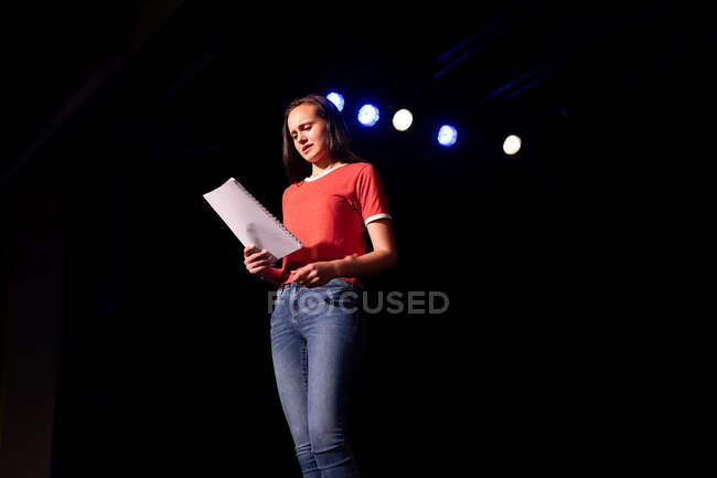 Vista frontale a basso angolo di una studentessa caucasica adolescente in un teatro vuoto della scuola superiore che si prepara prima di uno spettacolo, in piedi sul palco, con una sceneggiatura e pratica la sua parte — Foto stock