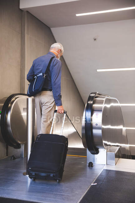 Senior Caucasian man, wearing a face mask against coronavirus, covid 19, entering an escalator in a metro station, pulling a suitcase. — Stock Photo
