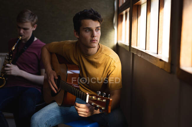 Вид спереди на белого подростка, сидящего у окна с акустической гитарой, а позади него сидит белый подросток, играющий на саксофоне в старшей школе — стоковое фото