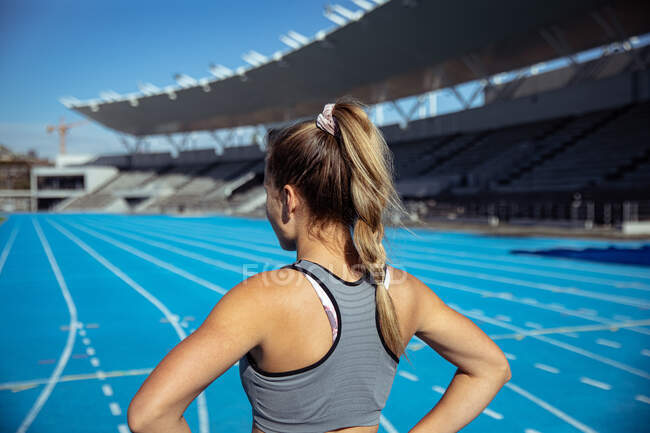 Vista posteriore di un'atleta caucasica con lunghi capelli biondi che pratica in uno stadio sportivo, concentrandosi prima dell'allenamento su una pista da corsa — Foto stock